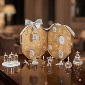 Swarovski Kristall Adventskalender 2026, geoeffnetes achteckiges Etui in Champagnerton mit davor aufgestellten Kristallobjekten, Adventskerzen und Kris Baer in weihnachtlichem Interieur