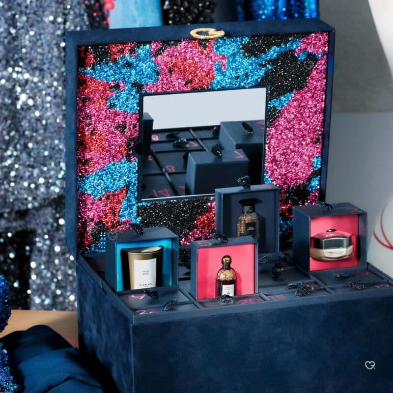 Guerlain Adventskalender 2026 geöffnet vor glitzerndem Hintergrund, mit Duftkerze und Parfums